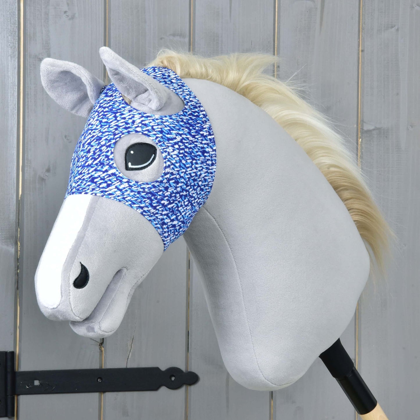 The Hobby Horse LarDen "Horse Mask without ears See" features a light gray face, white mane, blue-and-white patterned bridle, and a washable cover on a wooden stick, shown against a light wood panel background.