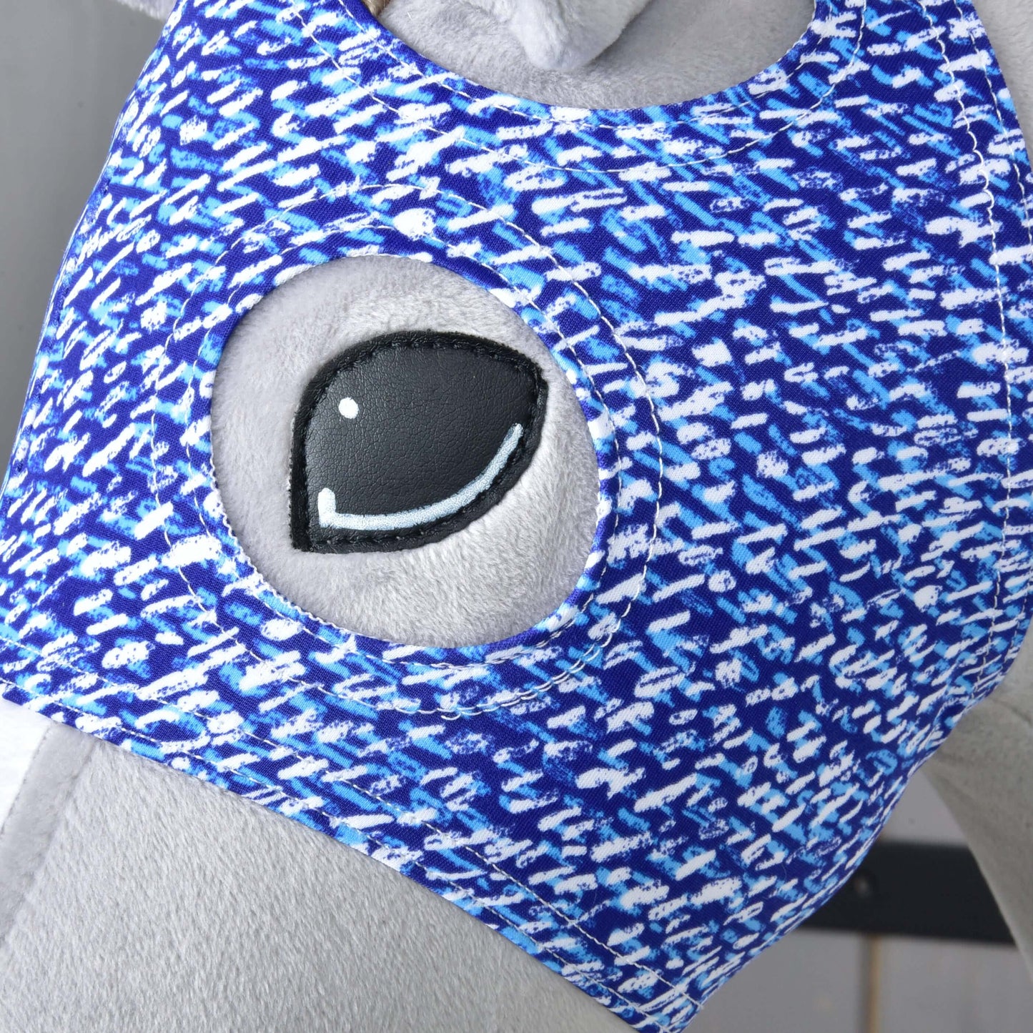 Close-up of a plush toy with grey fabric, featuring a large embroidered eye and wearing the Hobby Horse LarDen "Horse Mask without ears See," a blue and white washable cover with a circular opening for added hobby horse protection.