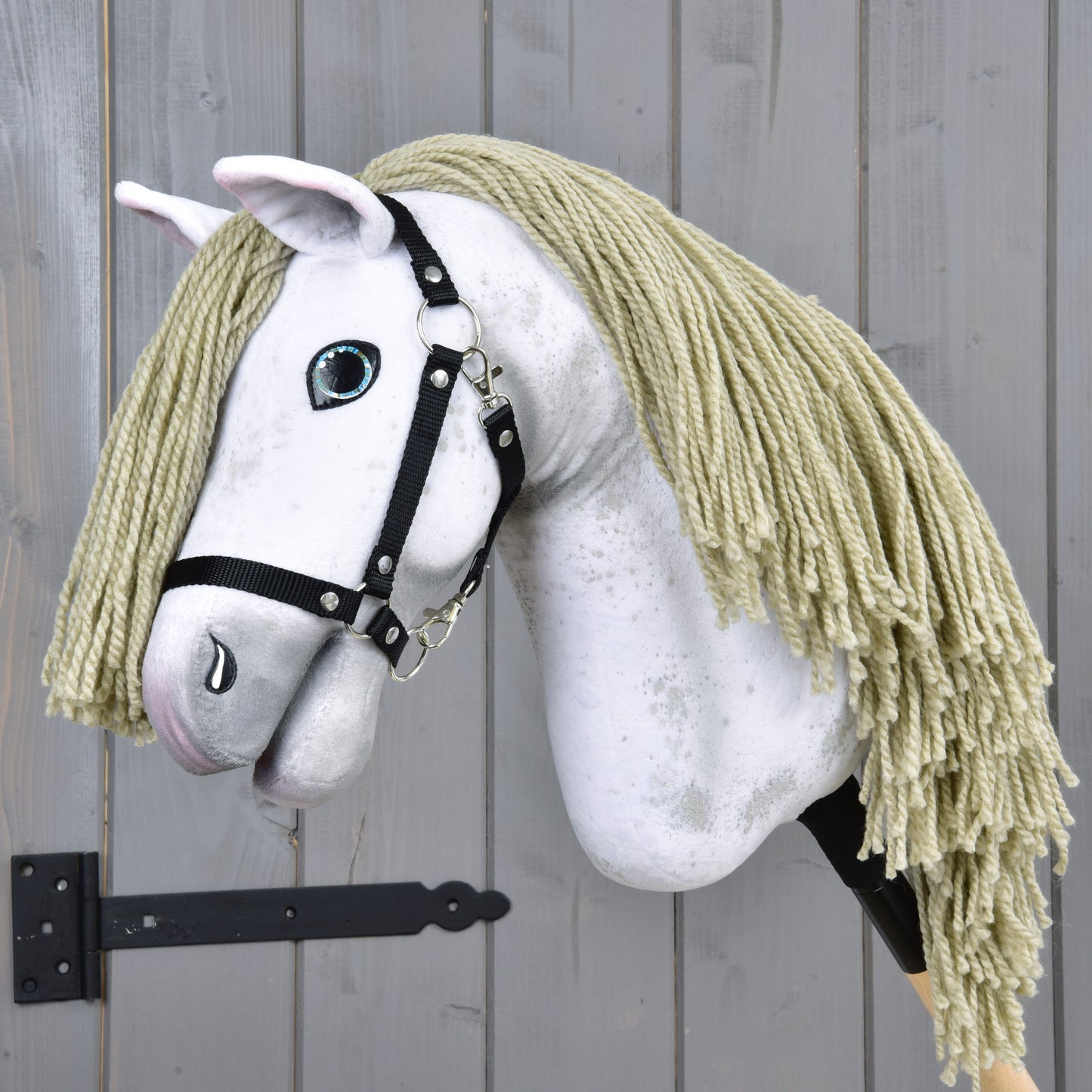 The Hobby Horse Victory with Black Halter - size M by Hobby Horse LarDen features a white plush head, blond yarn mane, black halter, and painted blue eye on a wooden stick, shown here before a gray wooden wall.