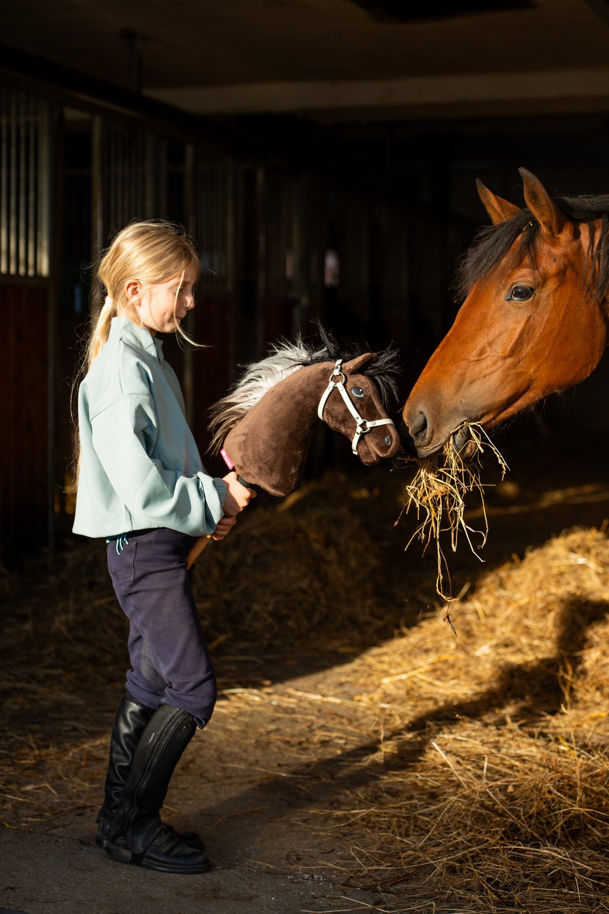Ragazza in maglia menta e stivali da equitazione in scuderia con Hobby Horse Alfredo in cavezza bianca accanto a un cavallo baio che mangia fieno