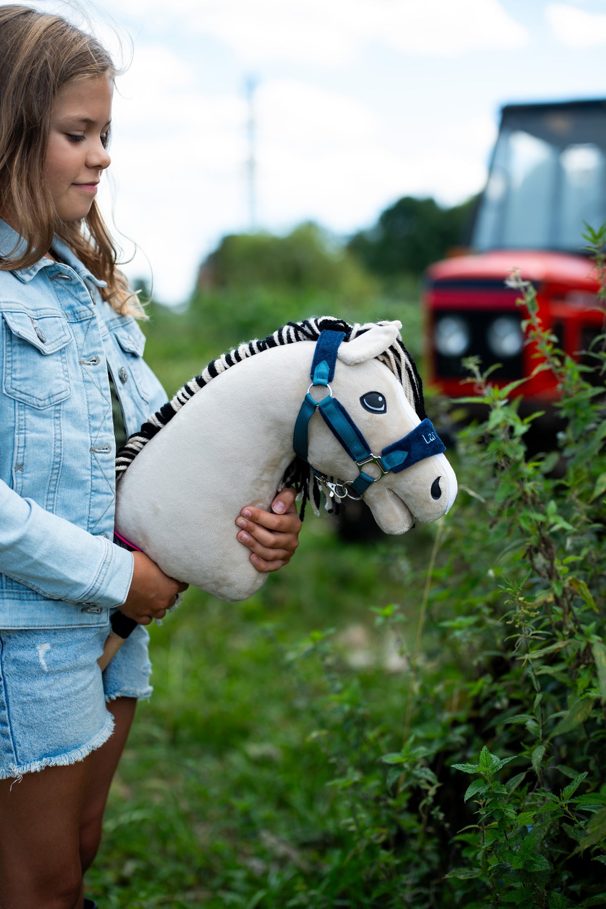 Ragazza con giacca di jeans accanto a una siepe con Hobby Horse Aurora con Cavezza nera di Hobby Horse LarDen, testa in peluche bianco crema e criniera in filato a righe nere e bianche, con Cavezza LarDen Blue foderata di pelliccia e trattore rosso sfocato sullo sfondo
