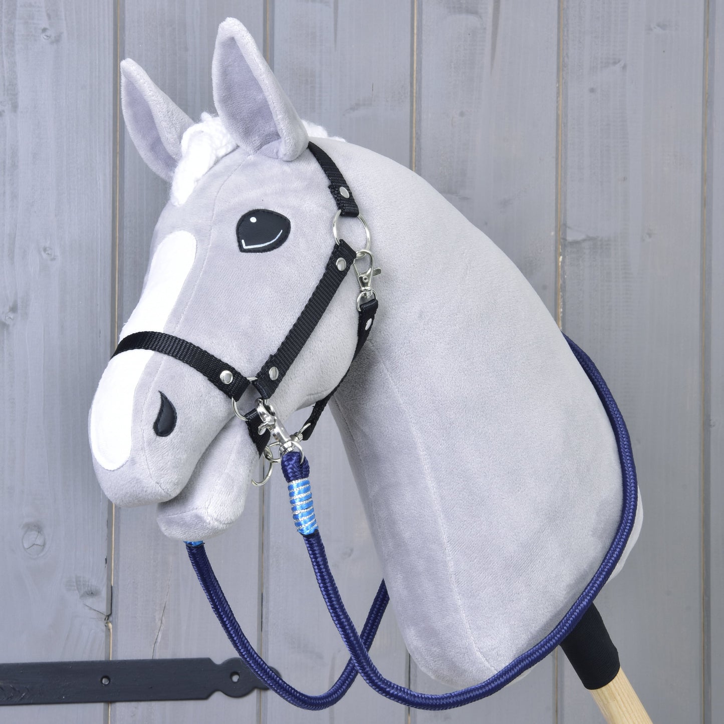 The Hobby Horse LarDen Reins Blues is a gray plush hobby horse with a black bridle, handmade reins, blue lead rope, and white face markings on a wooden stick, shown in front of a gray wooden wall.
