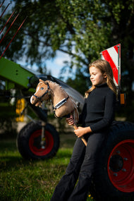 Ragazza con dolcevita nero accanto a macchinari agricoli verdi con Hobby Horse Bren con Cavezza nera di Hobby Horse LarDen, testa in peluche marrone sabbia e criniera in filato crema, con pettorale Espresso con pelliccia