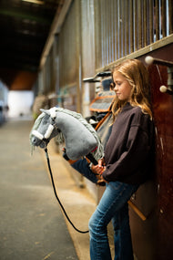 Ragazza con felpa marrone scuro appoggiata ai box nel corridoio di una scuderia con Hobby Horse Calypso con Cavezza nera di Hobby Horse LarDen, testa in peluche grigio e criniera in filato grigio chiaro, con Cavezza Teddy Classic foderata di pelliccia e Lunghina nera