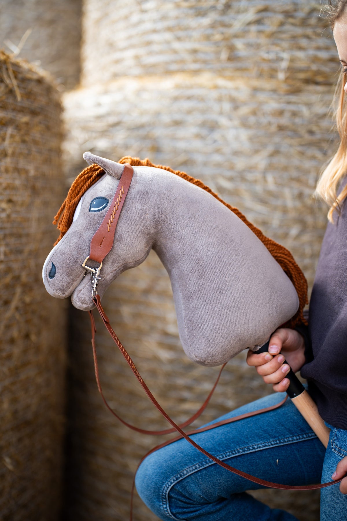 Detail photo: the head of the Hobby Horse Chanell with Black Halter by Hobby Horse LarDen held in front of round straw bales - grey plush, blue eye, ginger yarn mane - shown here with the complete Western one-ear bridle in brown leather.