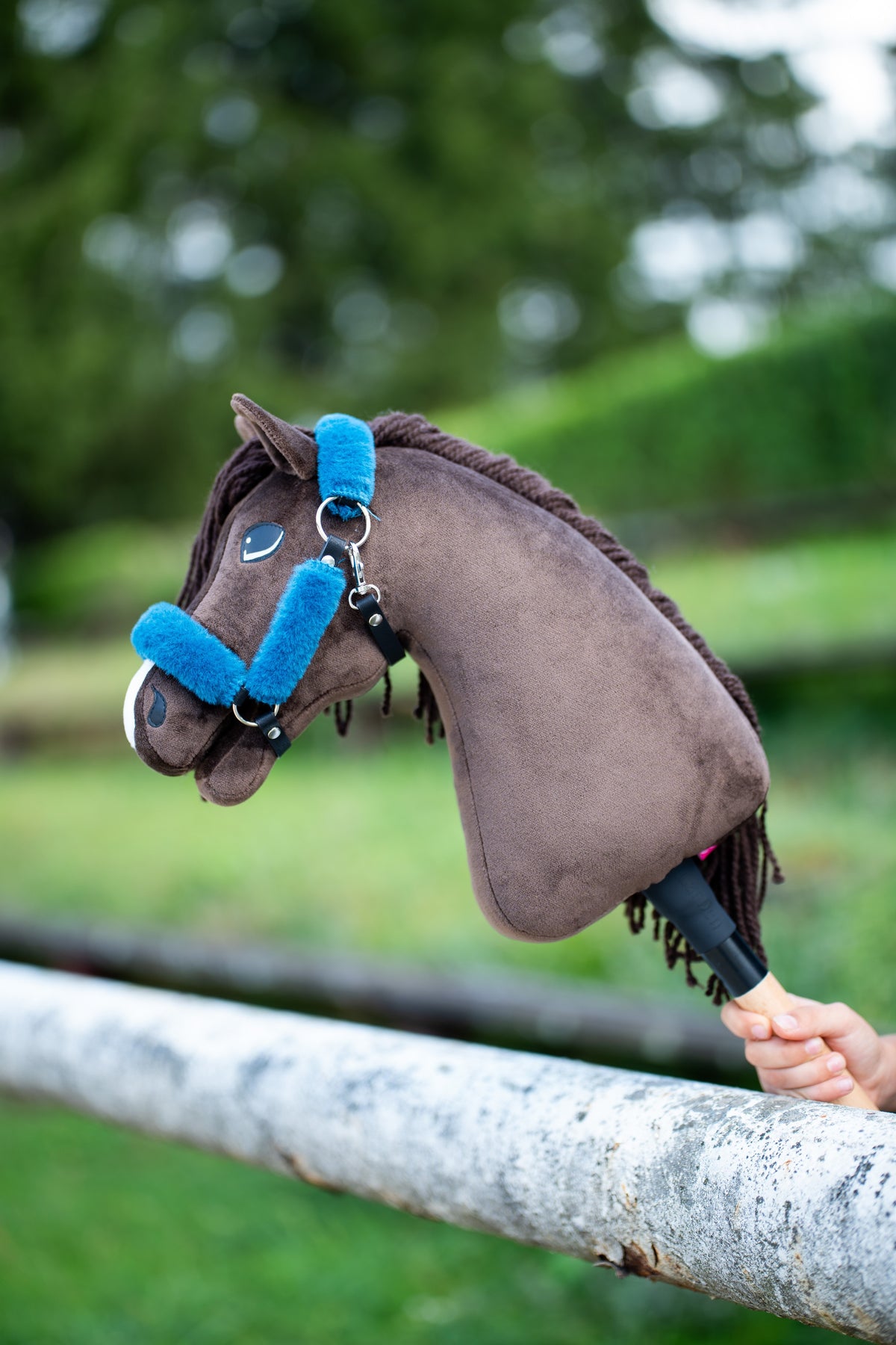 Hobby Horse Chianti con cavezza nera con pelliccia blu petrolio, appoggiato a una staccionata di betulla bianca davanti a cespugli verdi