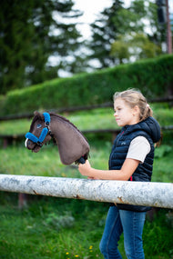 Ragazza con gilet imbottito blu navy dietro una staccionata bianca con Hobby Horse Chianti con Cavezza nera di Hobby Horse LarDen, testa in peluche marrone scuro e criniera in filato scuro, con Cavezza Teddy Petrol foderata di pelliccia e siepe verde potata sullo sfondo