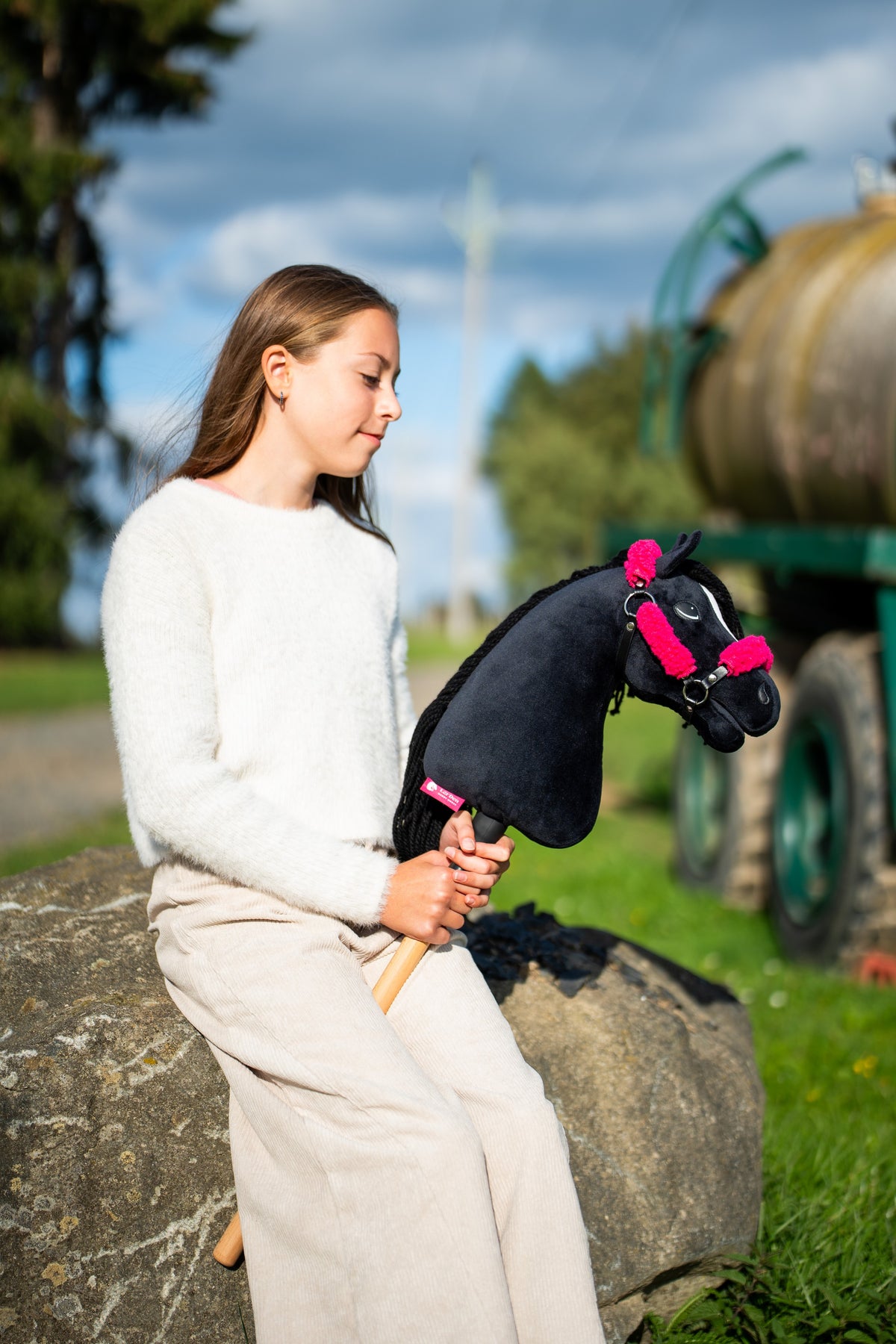 Ragazza con maglione crema seduta su un masso accanto a un sentiero nel prato con Hobby Horse Cloony con Cavezza nera di Hobby Horse LarDen, testa in peluche nero, stella bianca e criniera in filato nero, con Cavezza nera in similpelle con pelliccia rosa completa