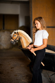 Ragazza con maglia bianca in una scuderia con Hobby Horse Colby con Cavezza nera di Hobby Horse LarDen, testa in peluche palomino, folta criniera in filato crema e Cavezza nera con finiture in argento