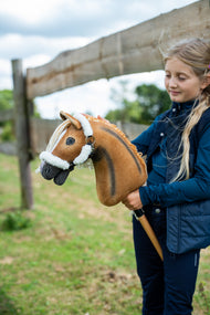 Ragazza con gilet blu navy accanto a una staccionata di legno con Hobby Horse Connie con Cavezza nera di Hobby Horse LarDen, testa in peluche sauro con striscia dorsale scura e criniera in filato crema, con Cavezza Teddy Classic foderata di pelliccia