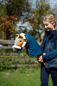 Lifestyle photo: a girl in a navy gilet holds the Hobby Horse Connie with Black Halter by Hobby Horse LarDen dressed in a petrol blue stable blanket, autumn berries behind - chestnut plush head, cream yarn mane - shown here with the fur-lined Teddy Classic halter and the Winter Sky stable blanket.
