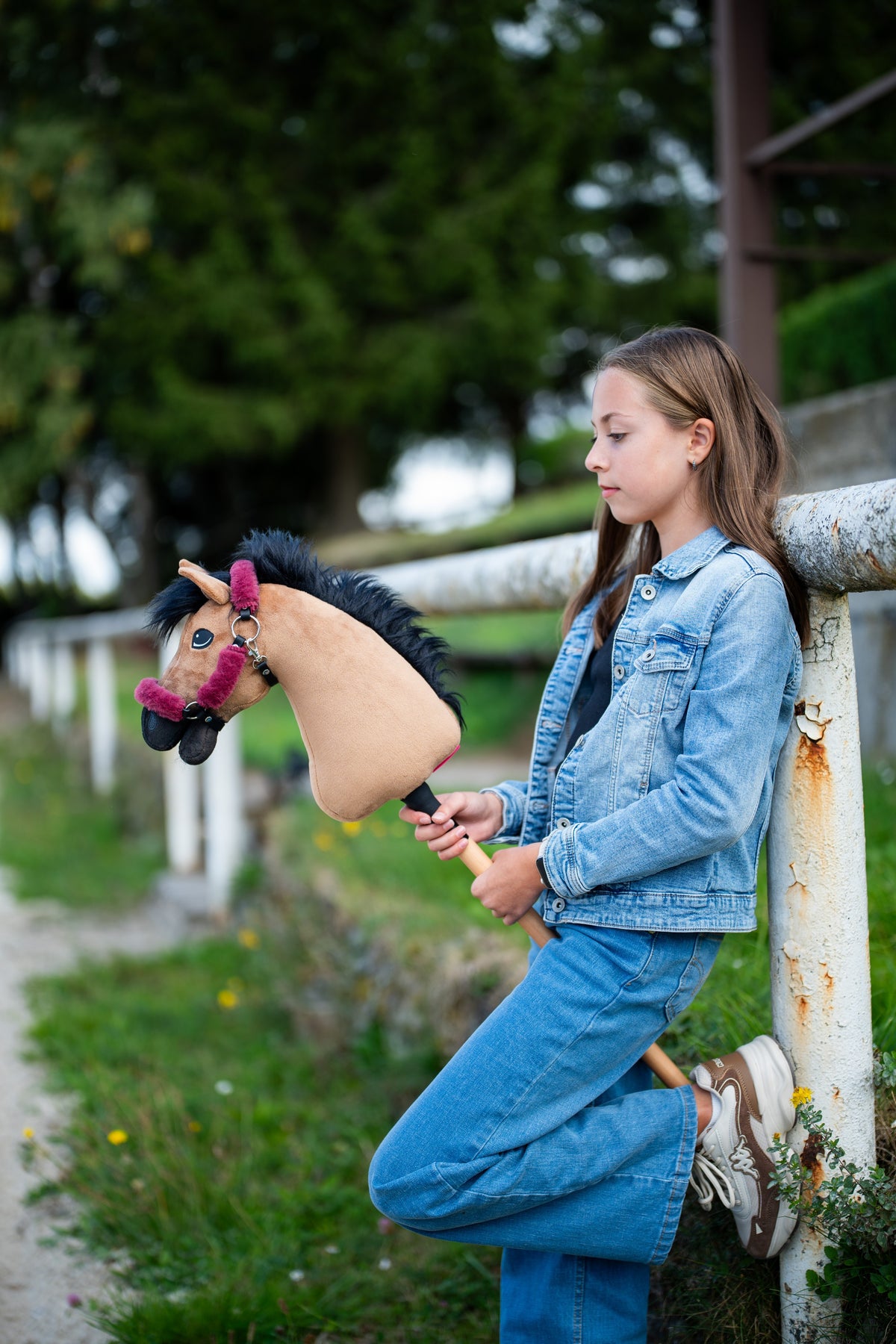 Ragazza in denim appoggiata a una staccionata bianca con Hobby Horse Cookie con Cavezza nera di Hobby Horse LarDen, testa in peluche color sabbia e criniera in filato nero, con Cavezza Teddy Maroon foderata di pelliccia