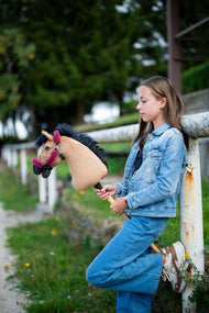 Ragazza in denim appoggiata a una staccionata bianca con Hobby Horse Cookie con Cavezza nera di Hobby Horse LarDen, testa in peluche color sabbia e criniera in filato nero, con Cavezza Teddy Maroon foderata di pelliccia
