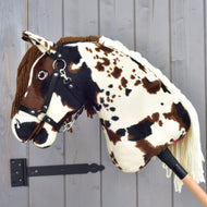 The Hobby Horse Dakota with Black Halter by Hobby Horse LarDen—a plush stick horse featuring brown and white spots, a bridle, and a yarn mane inspired by the wild Mustang horses of North America—is displayed against a gray wooden wall background.
