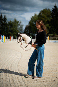 Ragazza con maglia nera in un campo in sabbia sotto un cielo tempestoso con Hobby Horse Frederik LD con Cavezza nera di Hobby Horse LarDen, testa in peluche crema e criniera in filato marrone scuro, con Briglia marrone completa con Nasello e ostacoli colorati sullo sfondo