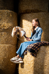 Ragazza con giacca di jeans seduta in alto su balle di paglia rotonde con Hobby Horse Fury con Cavezza nera di Hobby Horse LarDen, testa in peluche palomino con marcature bianche e criniera in filato crema, con Cavezza LarDen Brown foderata di pelliccia e Lunghina Bloom Brown