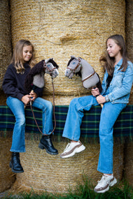 Due ragazze siedono su balle di paglia su una coperta tartan, ciascuna con un Hobby Horse Guccini di Hobby Horse LarDen dalla testa grigia in stile Holsteiner, lista bianca sul muso e lunga criniera in filato marrone scuro, con Cavezza Walnut foderata in pelliccia, pettorale Walnut con pelliccia e Lunghina Vivaldi
