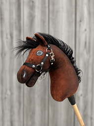 The Hobby Horse LarDen "Benny with Black Halter" (size XS) is a brown plush hobby horse with a black mane, wearing a black halter, shown on a wooden stick in front of a blurred wooden fence background.
