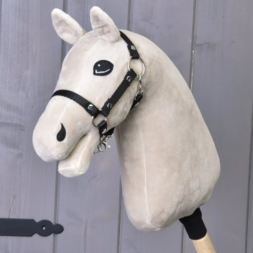 Costruisci il tuo cavallo, testa in peluche per Hobby Horse color beige chiaro con criniera bianca, briglia nera e montata su un bastone di legno, mostrata su sfondo di legno grigio.