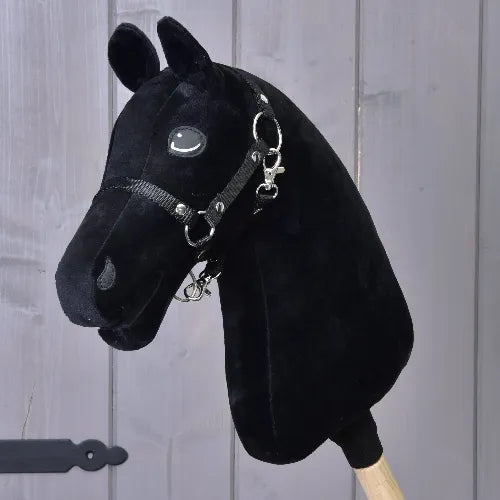 Costruisci il tuo cavallo, Hobby Horse con vistosa testa nera e occhi turchesi, completo di briglia e redini, montato su un bastone di legno e su sfondo di pannelli di legno grigi.