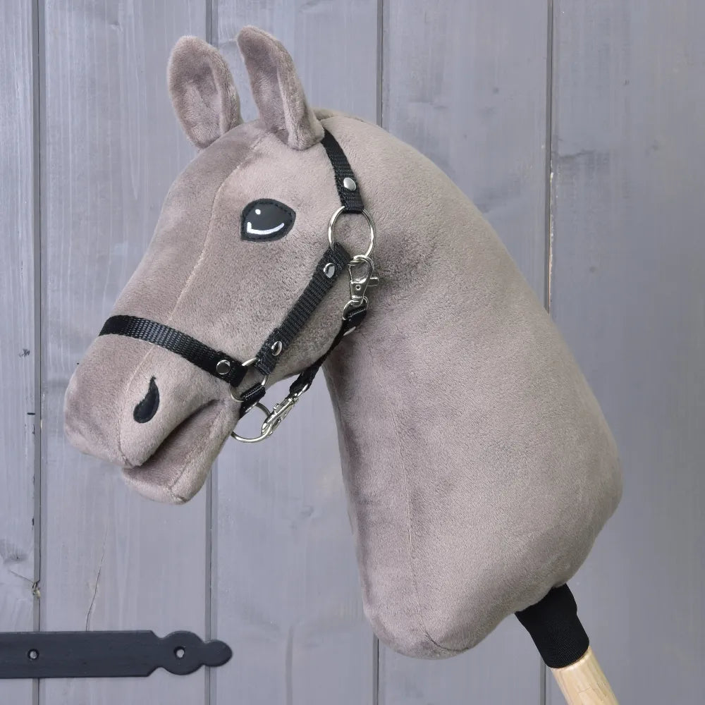 Costruisci il tuo cavallo, testa di Hobby Horse in peluche color tortora con occhi blu e briglia nera, montata su un bastone di legno e posizionata davanti a una parete di legno grigia.