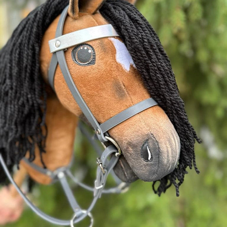 Primo piano di un Hobby Horse fatto a mano con criniera in filato nero, corpo marrone, una macchia bianca sopra un occhio e una briglia grigia, su sfondo esterno verde sfocato: un accessorio perfetto per le avventure di Hobby Horsing.