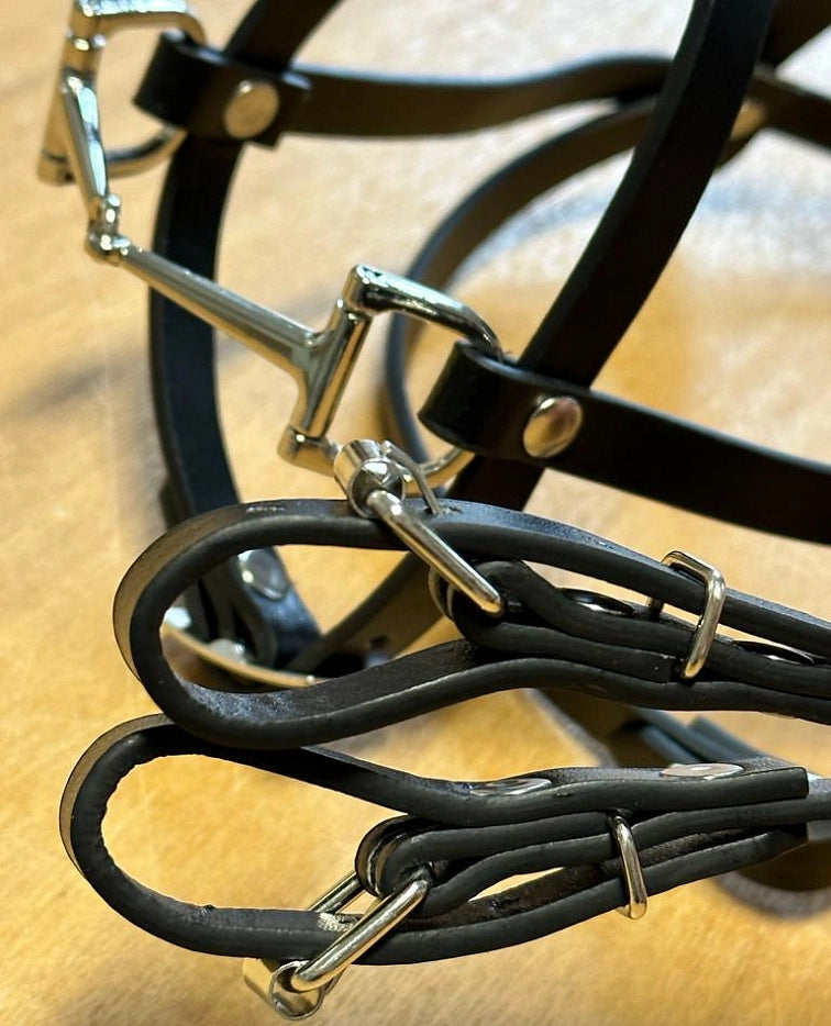 Primo piano di briglia e redini da cavallo in pelle nera con fibbie e anelli metallici argentati lucidi, perfette per una configurazione di Hobby Horsing, appoggiate su una superficie marrone chiaro.