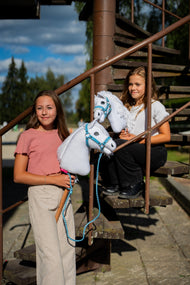 Due ragazze con Hobby Horses bianchi di Hobby Horse LarDen su una scala di metallo arrugginita, Hobby Horse Lady con Cavezza nera in primo piano, testa in peluche bianco e criniera in filato bianco, con Cavezza Pastell Blue con pelliccia in similpelle e Lunghina Double Blue lavorata a maglia