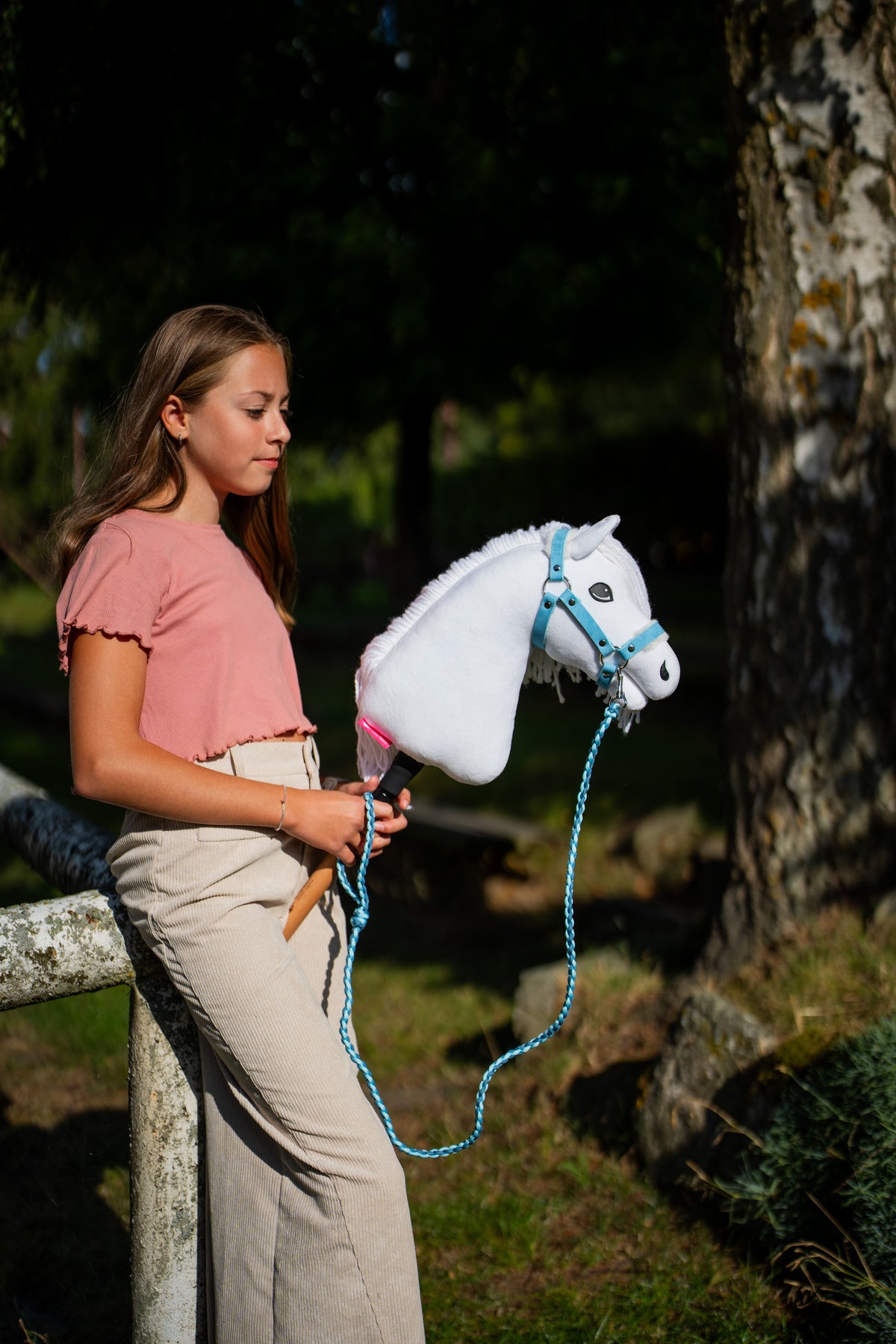 Ragazza con maglia rosa accanto a un albero muschioso con Hobby Horse Lady con Cavezza nera di Hobby Horse LarDen e Lunghina ad anello pendente, testa in peluche bianco e criniera in filato bianco, con Cavezza Pastell Blue con pelliccia in similpelle e Lunghina Double Blue lavorata a maglia
