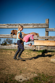Una ragazza in abbigliamento da equitazione attraversa un paddock sabbioso con l’Hobby Horse LarDen Pinkie con cavezza glitterata di Hobby Horse LarDen, testa in peluche rosa con criniera in lana rosa e bianca, qui con la Cavezza turchese
