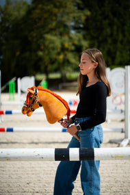 Ragazza con maglia nera dietro una barra di ostacolo in un campo in sabbia con Hobby Horse Little Angel con Cavezza nera di Hobby Horse LarDen, testa in peluche sauro e criniera in filato rosso acceso, con Cavezza Espresso con pelliccia in similpelle