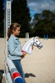 Ragazza con giacca di jeans accanto a un'ala di ostacolo in un campo in sabbia con Hobby Horse Little Lady con Cavezza nera di Hobby Horse LarDen, testa in peluche bianco e criniera in filato crema, con Cavezza grigia con pelliccia color menta