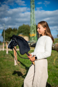 Una ragazza con maglione bianco sta in un prato soleggiato tenendo l'Hobby Horse Loki con Cavezza nera di Hobby Horse LarDen, testa in peluche nero, lista bianca e criniera in filato nero, qui mostrato con la briglia completa Pure Black senza nasello