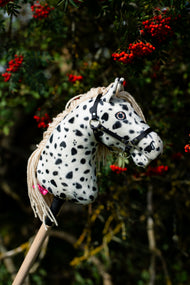 L'Hobby Horse Lord Valentine con Cavezza nera di Hobby Horse LarDen davanti a un sorbo con grappoli di bacche rosse, testa in peluche bianco con macchie leopardate nere, criniera in filato color crema e cavezza nera con finiture in argento