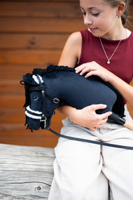 Una ragazza con top bordeaux tiene tra le braccia l'Hobby Horse Mahal con Cavezza nera di Hobby Horse LarDen davanti a una parete di legno, testa in peluche nero, criniera in filato nero e nasello in pelliccia bianca, qui mostrato con la briglia completa con pelliccia