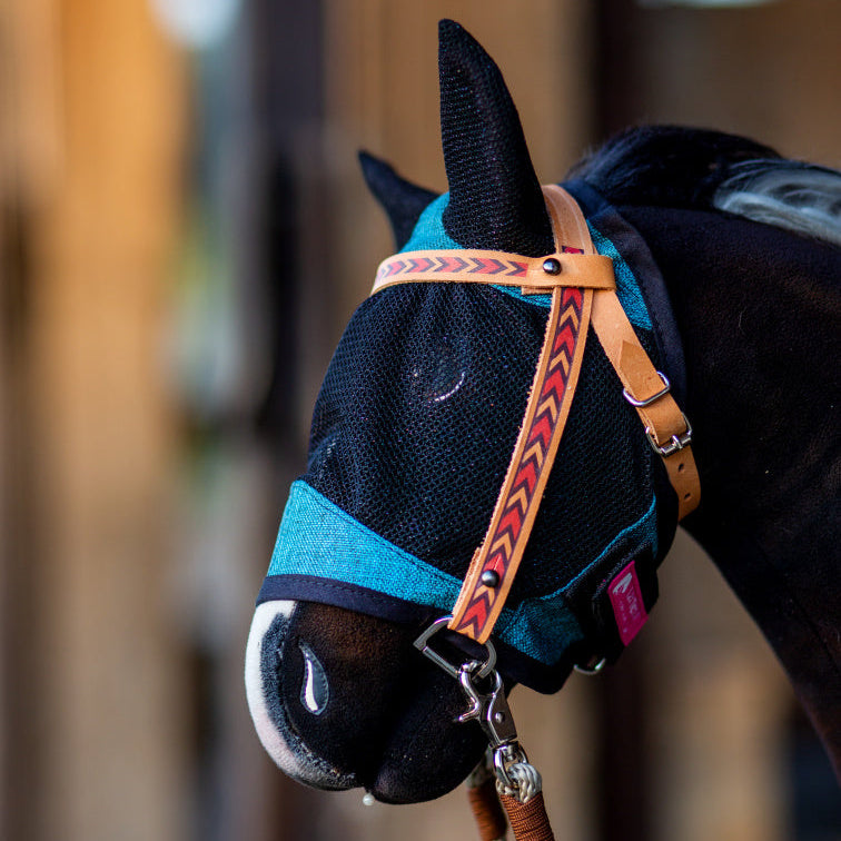 Primo piano di un cavallo che indossa una maschera antimosche in rete blu con copriorecchie e una cavezza in pelle marrone con motivo a freccia rosso, che ricorda l'attrezzatura per Hobby Horsing. Lo sfondo è leggermente sfocato.