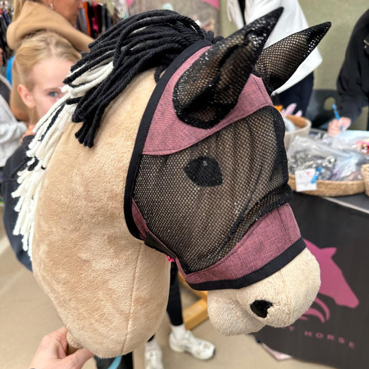 Primo piano di un Hobby Horse fatto a mano con maschera in rete e criniera in filato bianco e nero. Sullo sfondo sfocato, persone e uno stand con articoli a tema cavallo mostrano lo spirito creativo del Hobby Horsing.