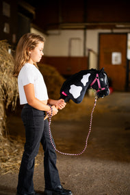 Una ragazza con un top bianco conduce l'Hobby Horse Merlin con Cavezza nera di Hobby Horse LarDen accanto a balle di paglia in una scuderia, con testa di peluche pezzata nera e bianca e criniera di filato nero, qui mostrato con il set LarDen Exclusive Pink