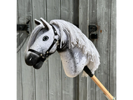 Primo piano di Hobby Horse Naivy con capezza nera di Hobby Horse LarDen, un Hobby Horse stallone Appaloosa grigio con criniera in filato bianco e macchie nere, posato davanti a una porta in legno invecchiata.
