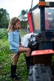 Una ragazza con giacca di jeans appoggia lo stivale sul gradino di un trattore rosso e nero, tenendo l'Hobby Horse Nexus con Cavezza nera di Hobby Horse LarDen, testa in peluche bianco e criniera in filato bianco, qui mostrato con la briglia completa De Luxe con due frontalini