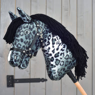 The Hobby Horse LarDen "Picasso" with Black Halter is styled as a striking stallion, featuring black and white leopard-patterned fabric reminiscent of an Appaloosa's spotted coat, complete with a black yarn mane and halter, all mounted on a wooden stick in front of a gray wooden door.
