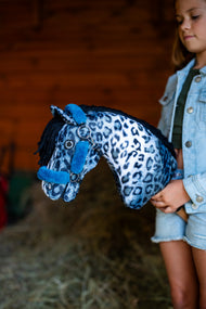 Una ragazza con giacca di jeans tiene l'Hobby Horse Picasso con Cavezza nera di Hobby Horse LarDen contro una parete rossa di legno del fienile, testa in peluche con stampa leopardata blu e bianca e criniera in filato nero, qui mostrato con cavezza Teddy Petrol foderata di pelliccia