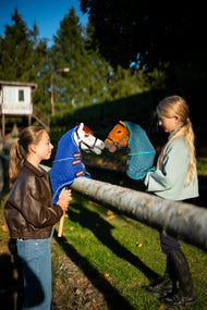 Lifestyle photo: two girls at a wooden rail hold the Hobby Horse Rufus with Black Halter in the Blanket with removable neck cover Bluebell and the Hobby Horse Pippa with Black Halter in the Blanket with removable neck cover Lagoon nose to nose in the evening sun - both by Hobby Horse LarDen.