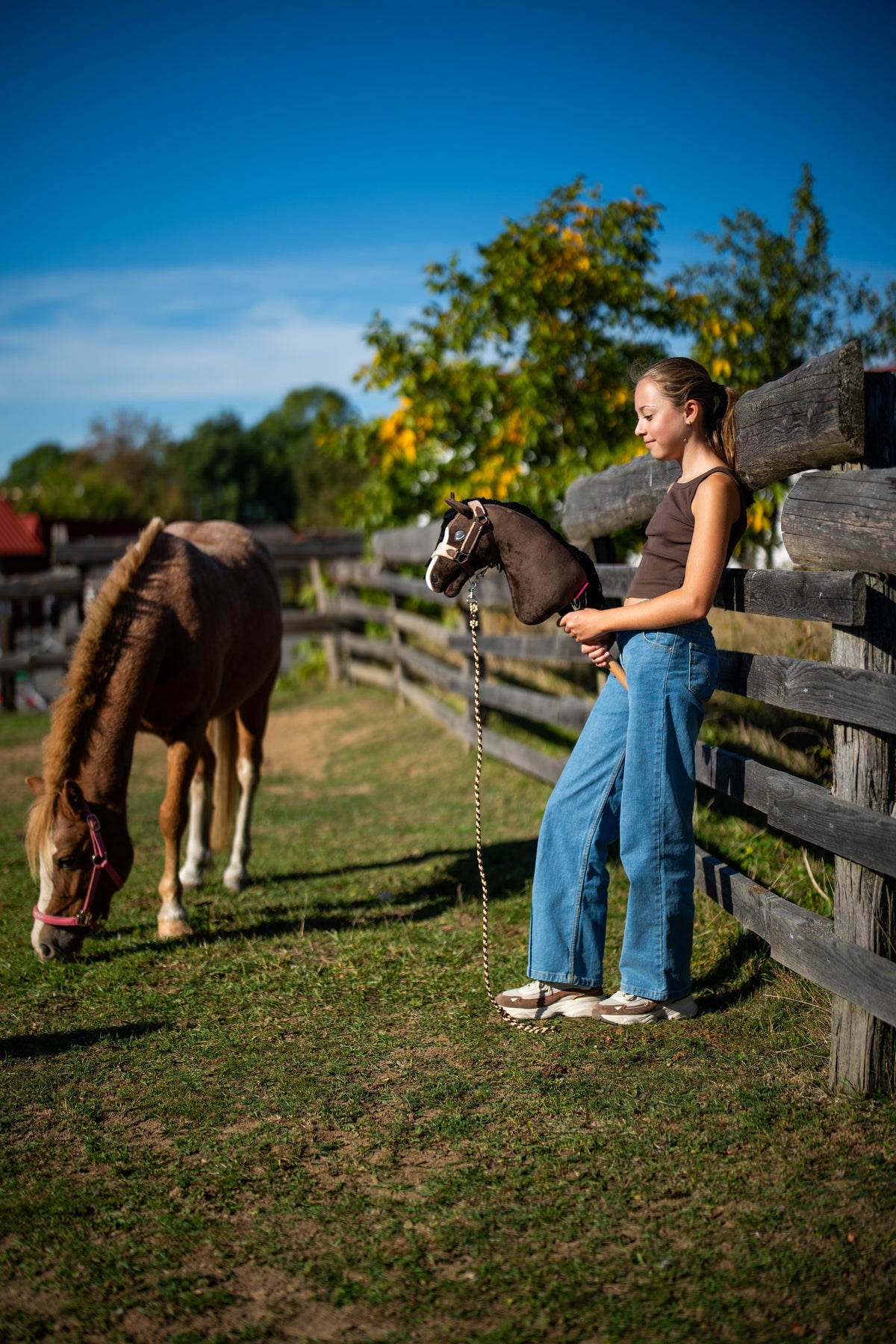 Ragazza in jeans alla staccionata del paddock tiene Hobby Horse Sisi alla lunghina mentre un pony sauro pascola vicino