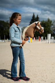 Una ragazza con giacca di jeans sta in un campo in sabbia tenendo l'Hobby Horse Smart con Cavezza nera di Hobby Horse LarDen, con ostacoli dietro, testa in peluche palomino e lunga criniera in filato color crema, qui mostrato con la cavezza nera in similpelle con pelliccia completa