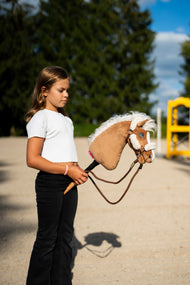 Una ragazza con top bianco porta l'Hobby Horse Star con cavezza nera di Hobby Horse LarDen attraverso un campo in sabbia, con un'ala gialla dell'ostacolo dietro, testa in peluche palomino e fluente criniera in filato bianco, qui mostrato con cavezza marrone con pelliccia completa e redini Vivaldi