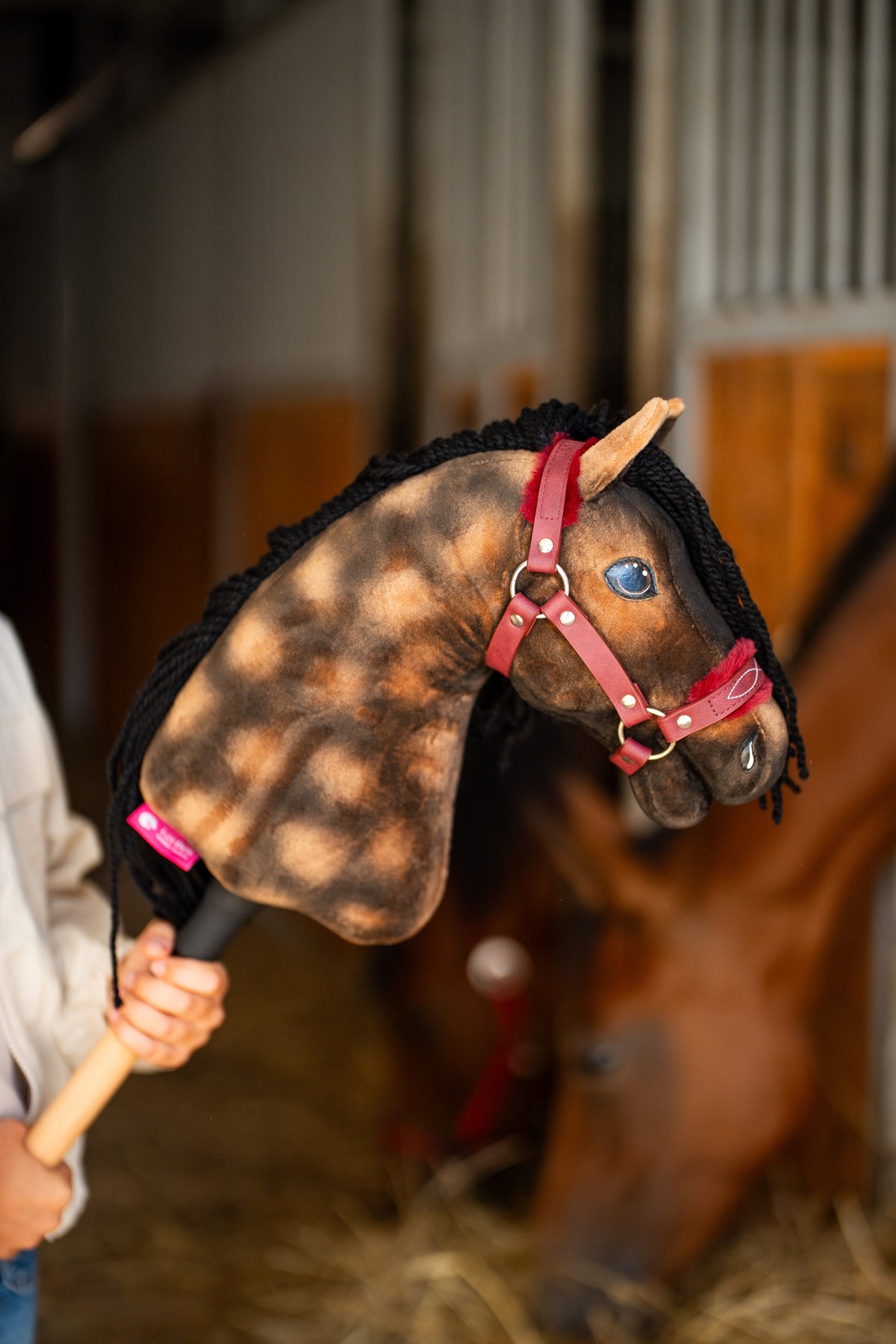 La testa dell'Hobby Horse Stormy con Cavezza nera di Hobby Horse LarDen, sollevato in una scuderia con un vero cavallo baio sfocato dietro, in peluche marrone pomellato con occhio blu e criniera in filato nero, qui mostrato con cavezza Burgundy in similpelle con pelliccia