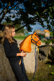Una ragazza con top nero si appoggia a un blocco di pietra sotto un albero tenendo l'Hobby Horse Wendy con Cavezza nera di Hobby Horse LarDen, testa in peluche sauro brillante e criniera in filato ramato, qui mostrato con lunghina Blues a righe in corda